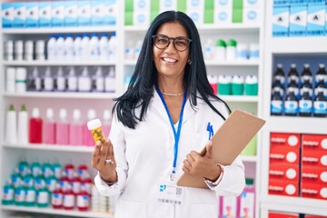Middle age hispanic woman pharmacist holding pills bottle reading document at pharmacy