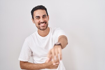 Handsome hispanic man standing over white background laughing at you, pointing finger to the camera with hand over body, shame expression