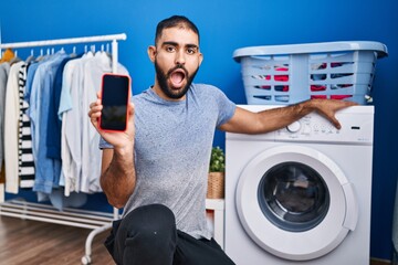 Middle east man with beard showing smartphone screen and washing machine in shock face, looking...