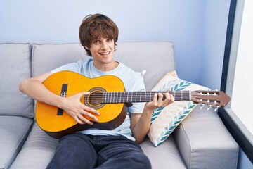 Young blond man playing classical guitar sitting on sofa at home