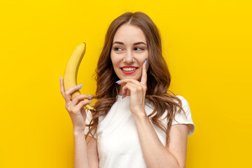 young pensive girl holding banana and smiling on yellow isolated background, woman dreams and...