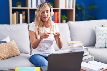 Young blonde woman studying using computer laptop at home pointing fingers to camera with happy and funny face. good energy and vibes.