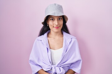 Young hispanic woman standing over pink background wearing hat with hands together and crossed...
