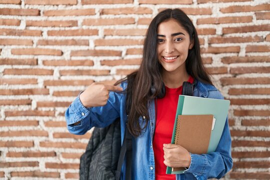 Young teenager girl wearing student backpack and holding books pointing finger to one self smiling happy and proud