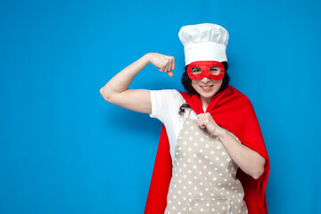 girl chef in superman costume shows strength on blue background, woman housewife in superhero mask...
