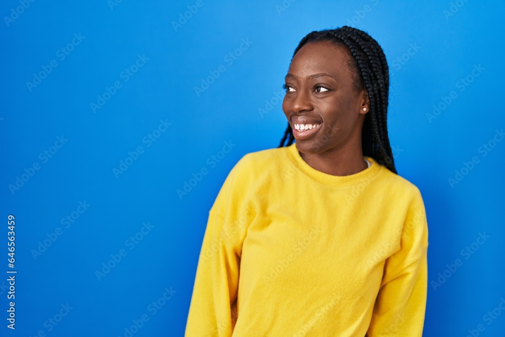 Wall mural Beautiful black woman standing over blue background looking away to side with smile on face, natural expression. laughing confident.