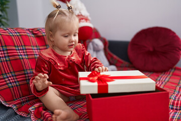 Naklejka premium Adorable blonde girl unpacking christmas gift sitting on sofa at home