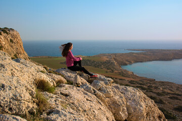 Woman sits on the edge of the cliff and looks into the distance at sunrise
