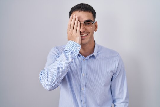 Handsome hispanic man wearing business clothes and glasses covering one eye with hand, confident smile on face and surprise emotion.