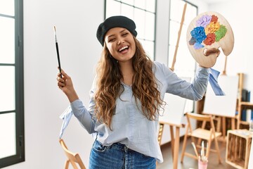 Young beautiful hispanic woman artist smiling confident holding paintbrush and palette at art studio