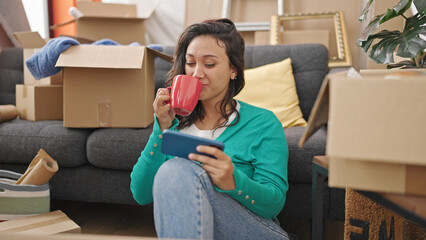 Young beautiful hispanic woman watching video on smartphone drinking coffee at new home
