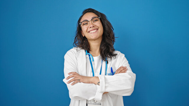 Young beautiful hispanic woman doctor smiling confident standing with arms crossed gesture over isolated blue wall background