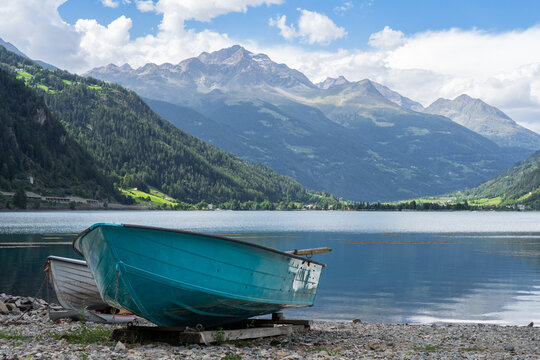 Swiss alpine lake in the mountains, turquoise boat in the foreground. Bernina massif peaks in the background, blue sky with clouds. Poschiavo (Miralago), Switzerland. 