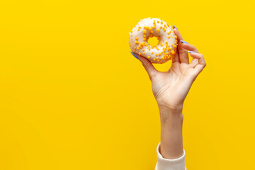 Fototapeta na wymiar hand raises and holds one sweet donut in white icing on yellow isolated background, girl shows and advertises dessert