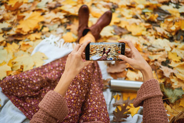 Top view of stylish autumnal woman taking photo of her legs on smartphone during picnic surrounded of orange leaves