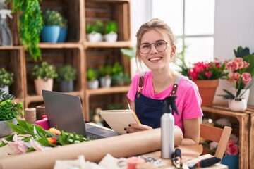 Young blonde woman florist using laptop writing on notebook at flower shop