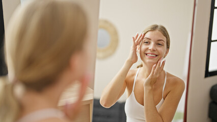 Young blonde woman touching face looking on mirror at home