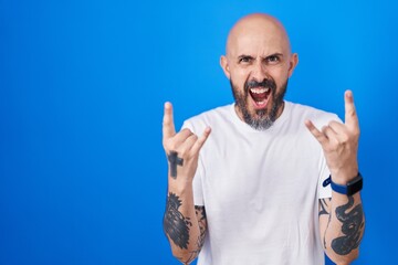 Naklejka premium Hispanic man with tattoos standing over blue background shouting with crazy expression doing rock symbol with hands up. music star. heavy music concept.