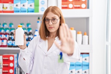 Young redhead woman working at pharmacy drugstore holding sun screen with open hand doing stop sign with serious and confident expression, defense gesture