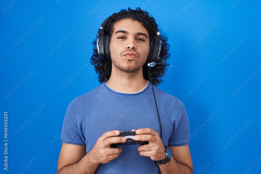 Poster hispanic man with curly hair playing video game holding controller looking at the camera blowing a k