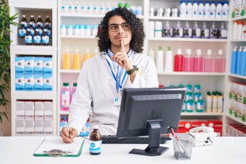 Hispanic man with curly hair working at pharmacy drugstore thinking concentrated about doubt with finger on chin and looking up wondering