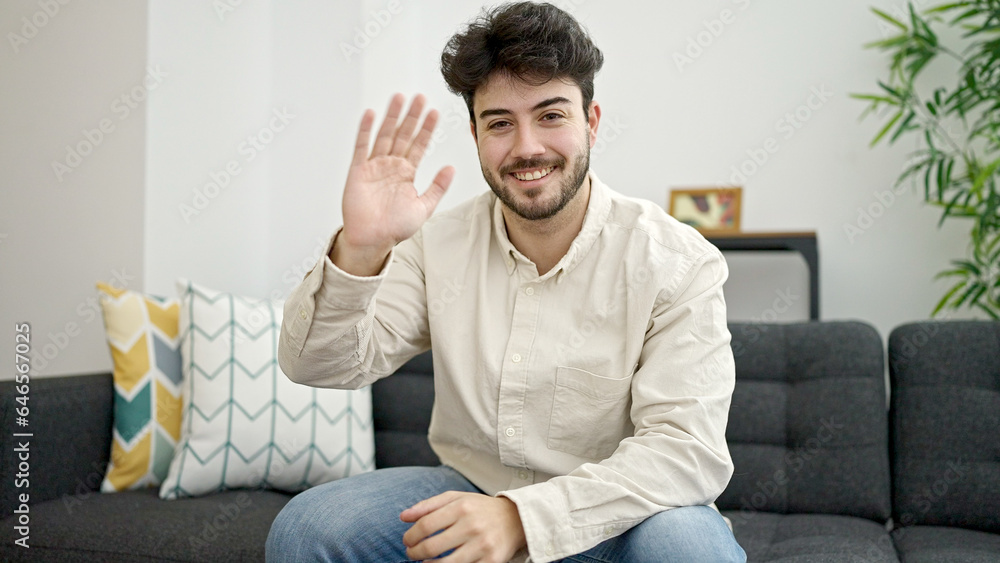 Sticker young hispanic man saying hello with hand sitting on sofa at home