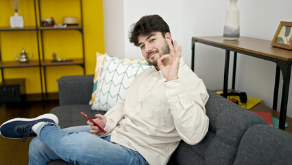 Young hispanic man using smartphone doing ok gesture at home