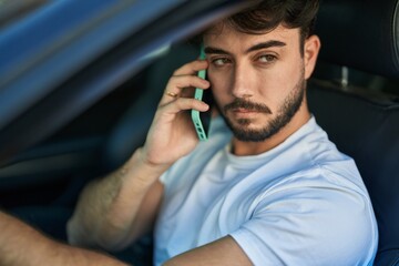 Young hispanic man talking on smartphone sitting on car at street