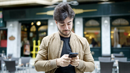 Young hispanic man using smartphone with serious expression at coffee shop terrace