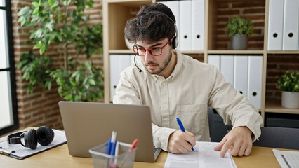 Young hispanic man call center agent writing notes speaking at office