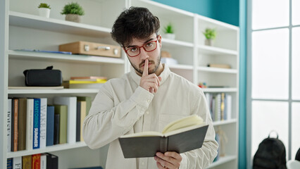 Fototapeta premium Young hispanic man student reading book doing silence gesture at library university
