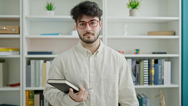 Young hispanic man student standing holding book at library university