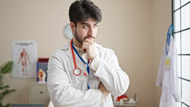 Young hispanic man doctor standing with doubt expression thinking at clinic
