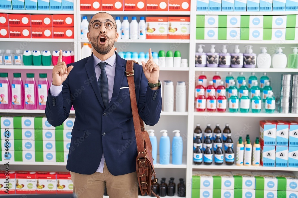 Poster Hispanic man with beard working as salesman at pharmacy drugstore amazed and surprised looking up and pointing with fingers and raised arms.