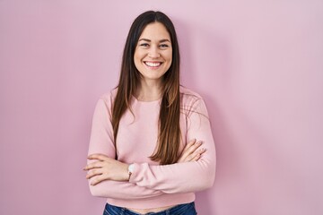 Young brunette woman standing over pink background happy face smiling with crossed arms looking at...