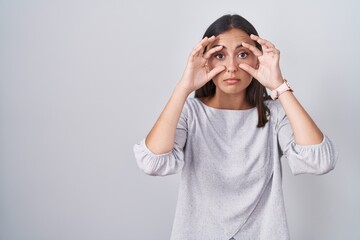 Young hispanic woman standing over white background trying to open eyes with fingers, sleepy and tired for morning fatigue
