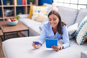 Young beautiful hispanic woman using touchpad and credit card lying on sofa at home