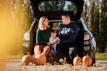 Family day. Happy mom, dad and little child daughter sit at car trunk, look at camera at autumn...