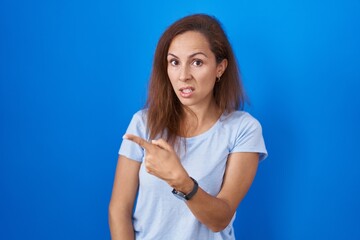 Brunette woman standing over blue background pointing aside worried and nervous with forefinger, concerned and surprised expression