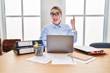 Young hispanic woman working at the office wearing glasses pointing finger up with successful idea. exited and happy. number one.