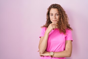 Obraz na płótnie Canvas Young caucasian woman standing over pink background looking confident at the camera smiling with crossed arms and hand raised on chin. thinking positive.