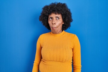 Fototapeta na wymiar Black woman with curly hair standing over blue background making fish face with lips, crazy and comical gesture. funny expression.