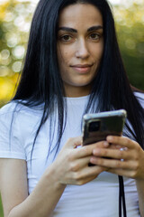 Close-up of a young woman with long dark hair holding a smartphone in her hands and browsing the news feed. Close-up of a woman's hand using a mobile phone outdoors. Girl in a white T-shirt