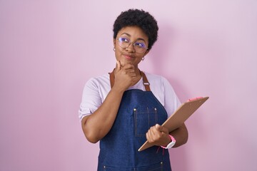 Young african american woman wearing professional waitress apron holding clipboard looking confident at the camera smiling with crossed arms and hand raised on chin. thinking positive.