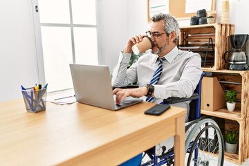Middle age grey-haired man business worker sitting on wheelchair drinking coffee at office