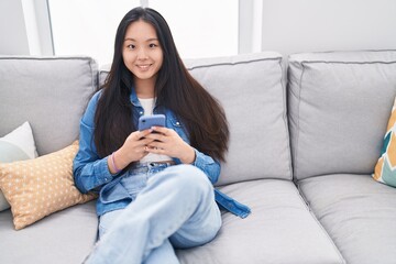 Young chinese woman using smartphone sitting on sofa at home