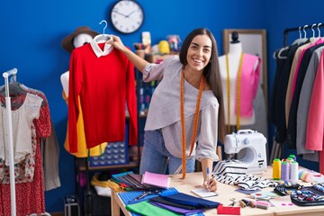 Young beautiful hispanic woman tailor smiling confident holding t shirt writing on notebook at clothing factory