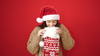 Young beautiful hispanic woman wearing christmas hat looking inside of sock over isolated red background