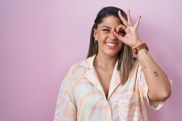 Blonde woman standing over pink background doing ok gesture with hand smiling, eye looking through fingers with happy face.