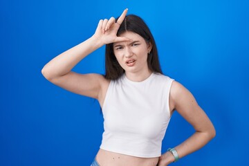Fototapeta na wymiar Young caucasian woman standing over blue background making fun of people with fingers on forehead doing loser gesture mocking and insulting.